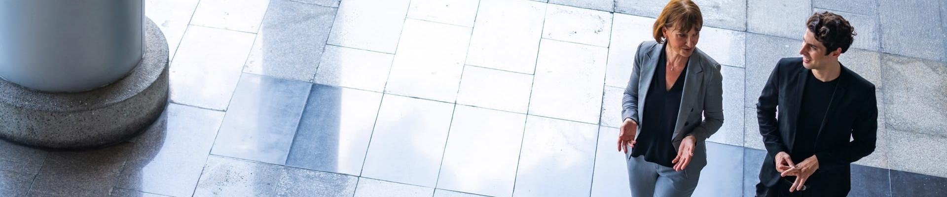 Frau und Mann in Businesskleidung laufen gemeinsam durch eine moderne, mit Marmor ausgekleidete, Halle