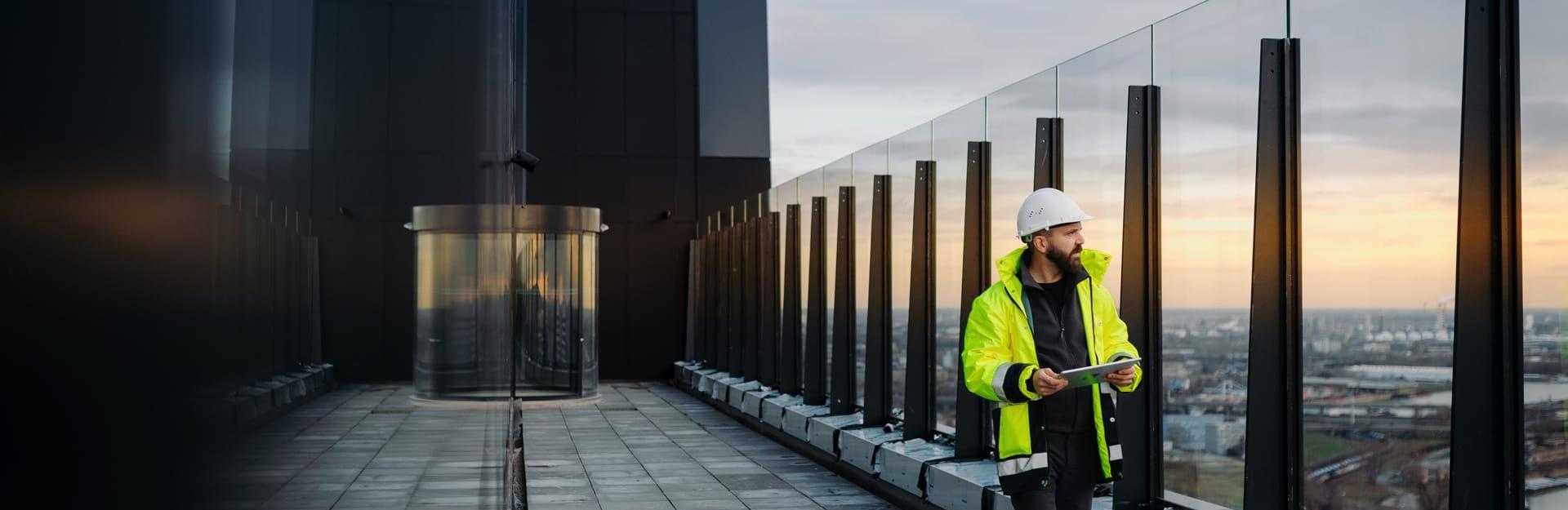 Ein Bauingenieur in Sicherheitskleidung überprüft die Fassade eines Hochhauses mit Tablet bei Sonnenuntergang – moderne Gebäudetechnik und nachhaltige Architektur.