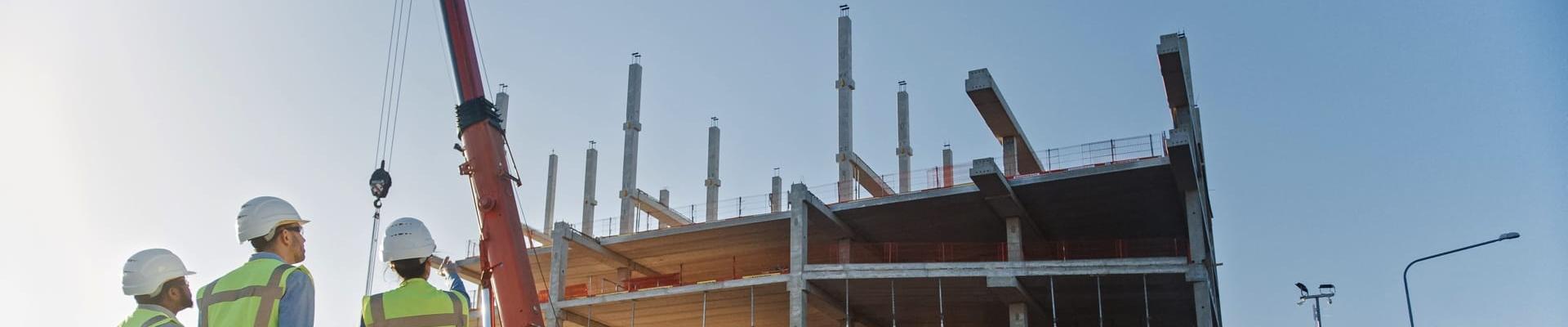 Drei Personen in Sicherheitskleidung und mit Helmen auf einer Baustelle im Gespräch mit Blick auf Baufahrzeuge und Baustelle